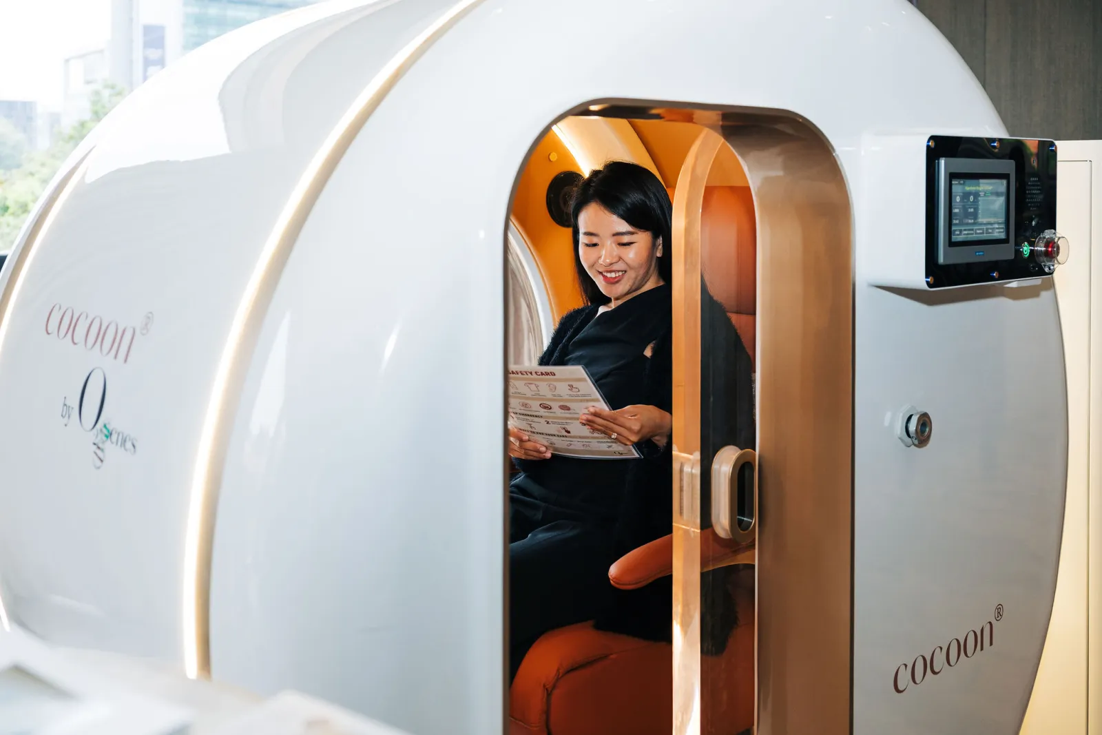 A visitor seated inside an O2genes Hard Shell HBOT cocoon® reading the product safety card